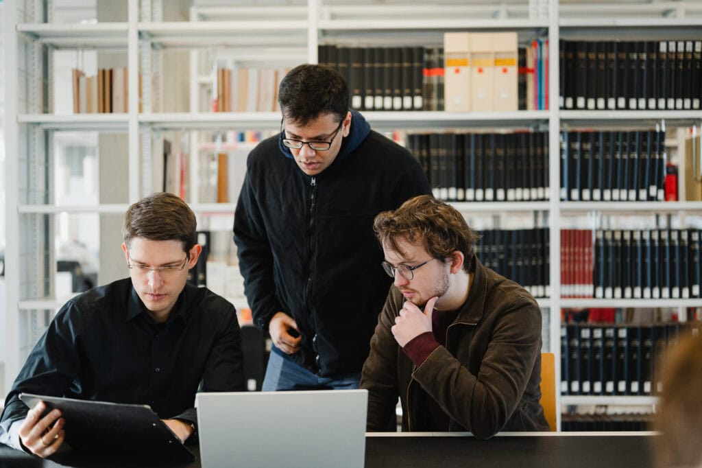 AI made in Germany: Ein zukunftsweisender Bildpool für die Konrad Zuse Schools (DAAD) 12 Drei junge Männer arbeiten in einer Bibliothek an einem Laptop.