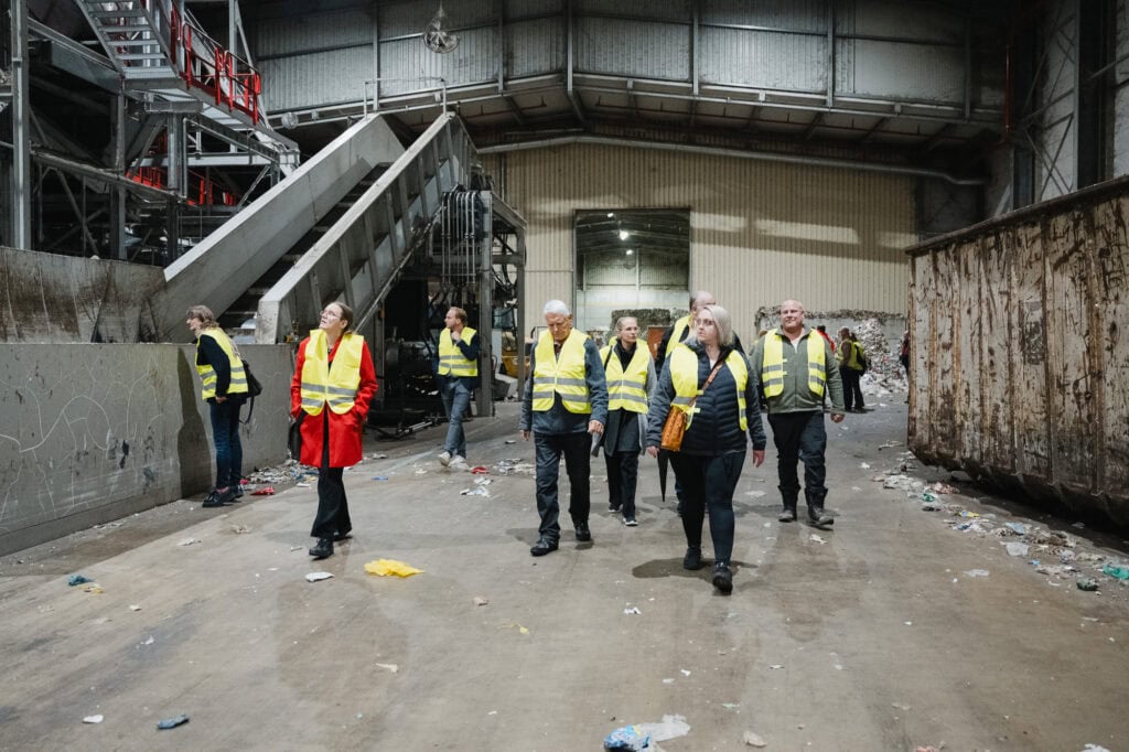 Gruppen von Menschen in Sicherheitswesten bei einer Werksbesichtigung in einer Industriehalle.